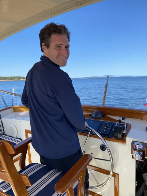 Sam at the helm of a boat on a bright day in Maine, looking back over his shoulder