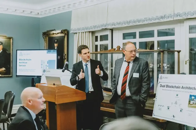 Sam, two thumbs up, presenting beside a technology founder at an ALPS executive event, with a blockchain architecture poster behind them
