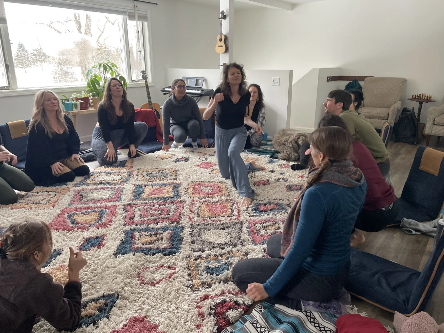 A circle on a patterned rug in a bright room, snow outside, one person standing to speak