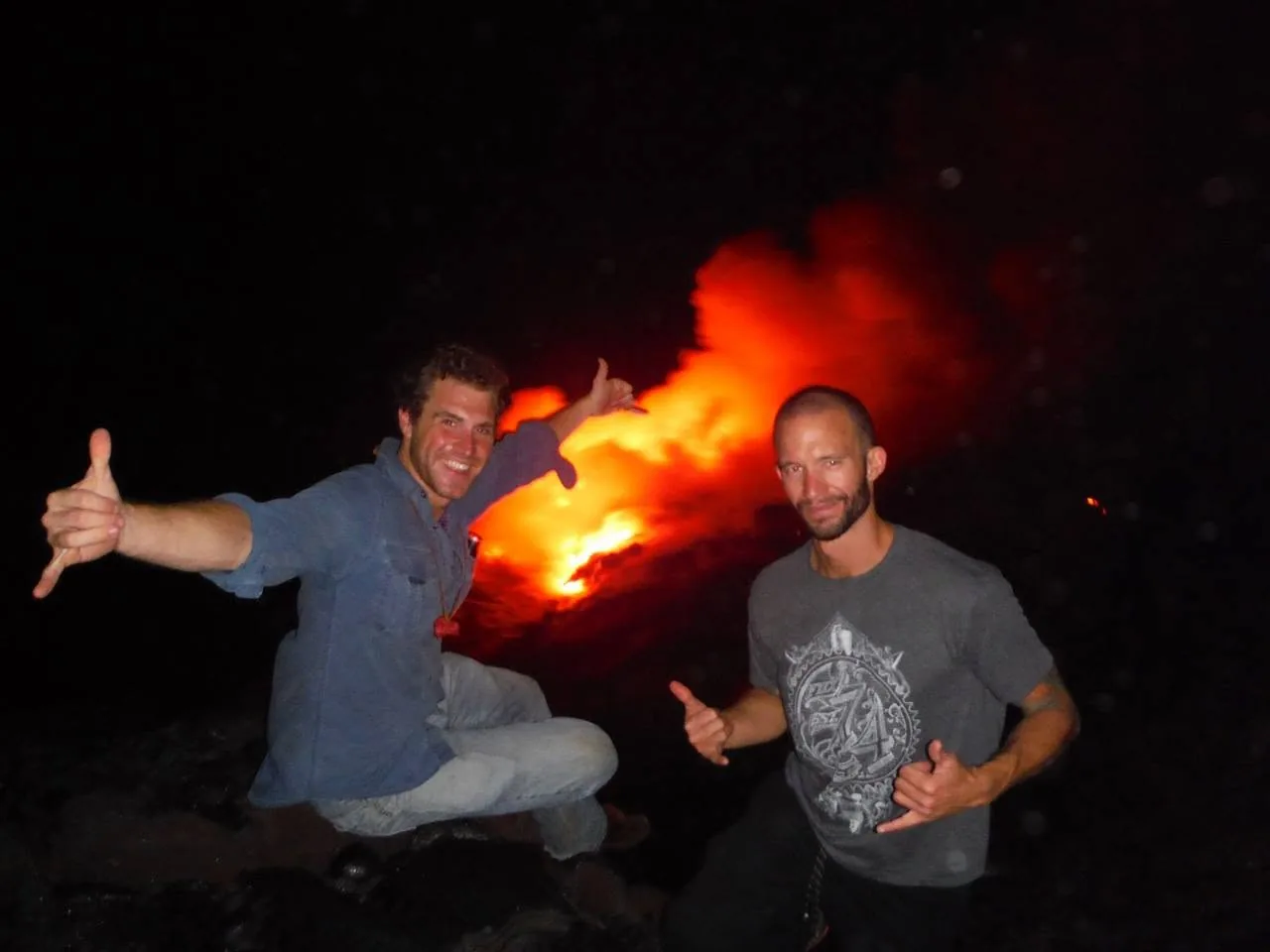Two men throwing shakas at night in front of glowing red lava on the Big Island of Hawaiʻi
