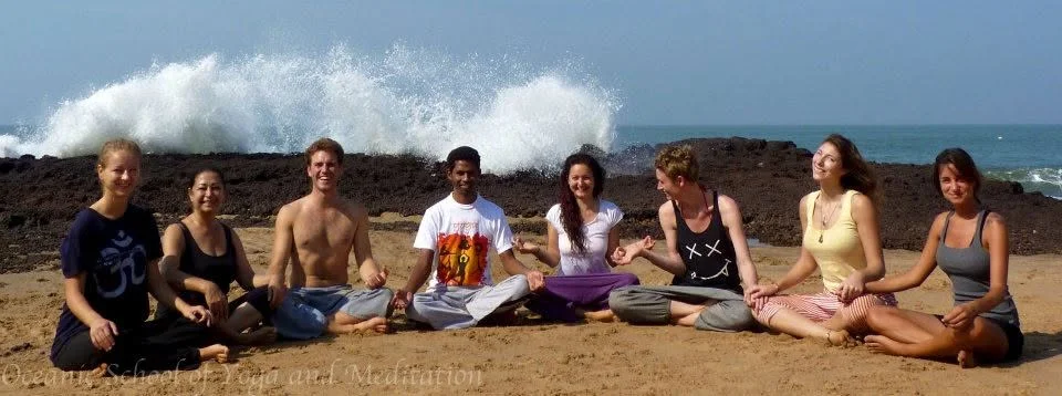 A yoga group seated on the rocks at the shore as a wave breaks behind them