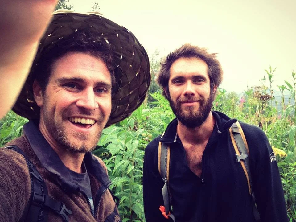 Two travellers in wide-brimmed hats, grinning, on an overgrown trail