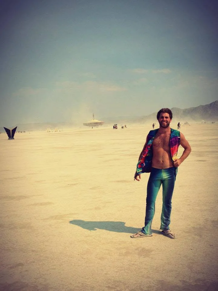 A faded print: shirtless in jeans on a wide white beach, a shirt over one shoulder